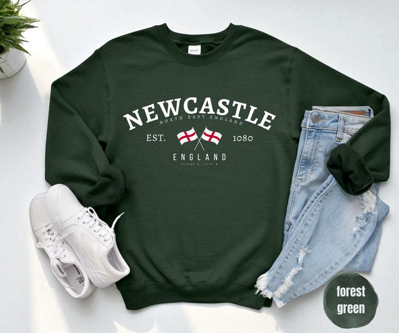 Puede incluir: Sudadera verde oscuro con la palabra "Newcastle" en blanco. Debajo, dos banderas cruzadas y la palabra "England". Zapatillas blancas y vaqueros azules junto a la sudadera.