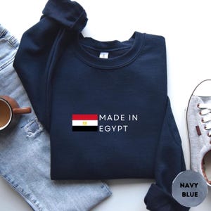 May include: Navy blue sweatshirt with the text "MADE IN EGYPT" and the Egyptian flag. The sweatshirt is folded and placed on a light-colored surface, next to a pair of jeans, a mug, and a pair of sneakers.