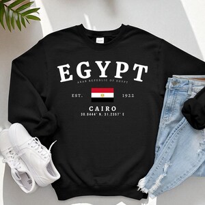 May include: Black sweatshirt with the word "EGYPT" in white block letters above a flag of Egypt. Below the flag are the words "CAIRO" and coordinates. The sweatshirt is paired with light blue jeans and white sneakers.