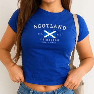 May include: A royal blue crop top featuring the word "SCOTLAND" in white, with a white and blue Scottish flag graphic. Below the flag is the city name "EDINBURGH" and coordinates. The shirt has short sleeves and a crew neck.
