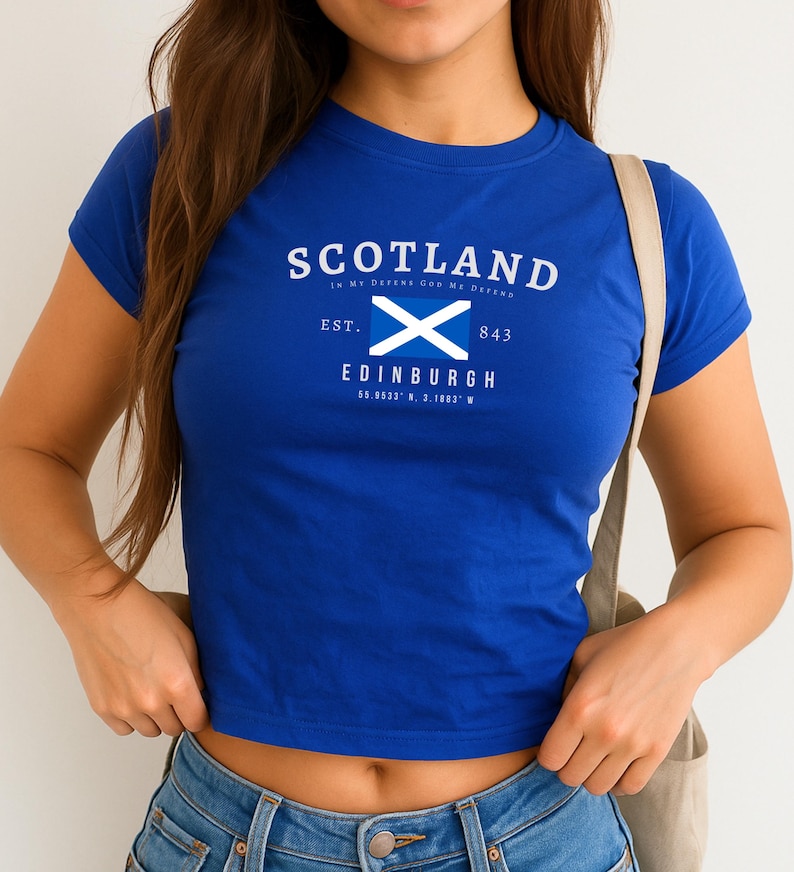May include: A royal blue crop top featuring the word "SCOTLAND" in white, with a white and blue Scottish flag graphic. Below the flag is the city name "EDINBURGH" and coordinates. The shirt has short sleeves and a crew neck.