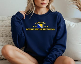 Könnte beinhalten: Ein marineblaues Sweatshirt mit dem gelben Schriftzug "Sarajevo", der sich über den Worten "Bosnien und Herzegowina" wölbt. Das Design beinhaltet die bosnische Flagge und das Jahr "1992". Das Sweatshirt hat einen Rundhalsausschnitt.