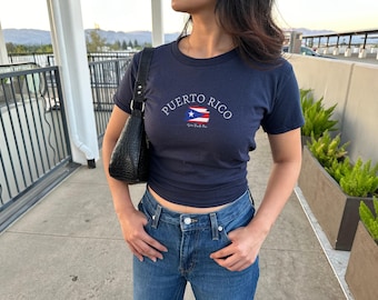 May include: A navy blue t-shirt with a white graphic of the Puerto Rican flag and the text "PUERTO RICO" with the words "Viva Puerto Rico" underneath. The shirt is being worn by a person with long dark hair and is paired with blue jeans.