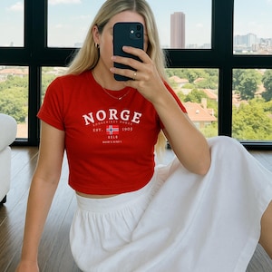 以下が含まれることがあります： ノルウェーの国旗と「NORGE」、「KONGERIKET NORGE」、「EST. 1905」、「BELO」の文字が白でプリントされた赤いクロップドTシャツ。