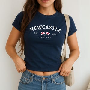 May include: Navy blue t-shirt with the word "Newcastle" in white arched text. Below the text are two crossed flags and the words "North East England" and "England". The shirt also has the text "Est. 1080".