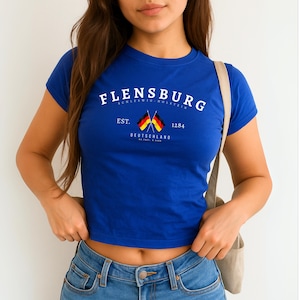 Puede incluir: Una camiseta corta azul real con la palabra "FLENSBURG" en texto blanco arqueado. Debajo, dos banderas alemanas cruzadas, el texto "EST. 1284" y "DEUTSCHLAND". La camiseta se combina con unos vaqueros azules.