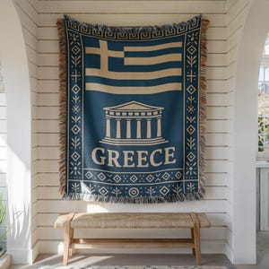 Könnte beinhalten: Eine blau-beige gewebte Decke mit der griechischen Flagge und dem Wort "GREECE". Die Decke hat einen Fransenrand und hängt an einer weißen Wand über einer Holzbank.