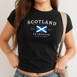 May include: Black t-shirt with the word "SCOTLAND" in white, along with the phrase "IN MY DEFENSE GOD ME DEFEND". Below is a blue and white flag, the word "EDINBURGH", and coordinates. The shirt is worn with blue jeans.