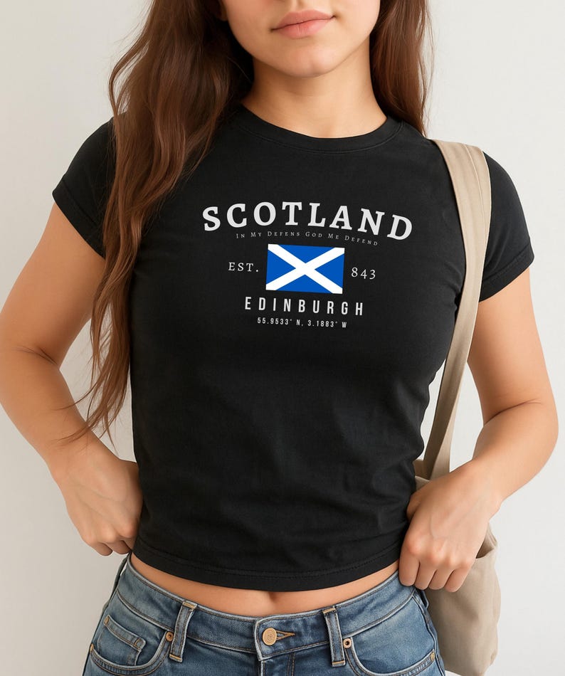 May include: Black t-shirt with the word "SCOTLAND" in white, along with the phrase "IN MY DEFENSE GOD ME DEFEND". Below is a blue and white flag, the word "EDINBURGH", and coordinates. The shirt is worn with blue jeans.