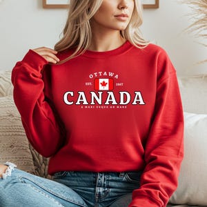 Pode incluir: Um moletom vermelho com a palavra "CANADA" em letras brancas. Acima da palavra "CANADA" está a palavra "OTTAWA" e uma bandeira canadense. O moletom também tem o texto "A MARI USQUE AD MARE" abaixo.
