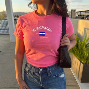Może przedstawiać: R&oacute;żowy T-shirt z białym nadrukiem flagi Salwadoru i tekstem "El Salvador Est. 1980 El Salvador"