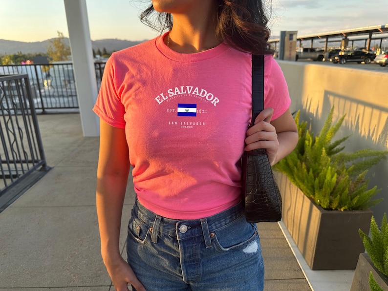 Może przedstawiać: R&oacute;żowy T-shirt z białym nadrukiem flagi Salwadoru i tekstem "El Salvador Est. 1980 El Salvador"