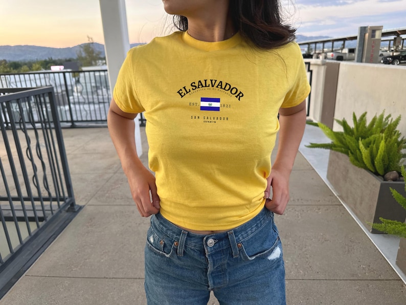 Może przedstawiać: Ż&oacute;łta koszulka z napisem "EL SALVADOR" i małym graficznym przedstawieniem niebiesko-białej flagi. Tekst pod flagą brzmi "EST. 1921 SAN SALVADOR"