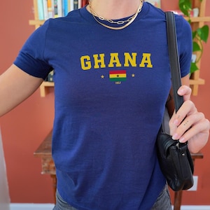 K&ouml;nnte beinhalten: Ein marineblaues T-Shirt mit dem Wort "GHANA" in gelben Buchstaben und einer kleinen ghanaischen Flagge mit dem Jahr "1957" darunter.