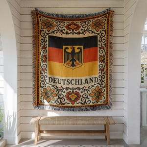 May include: A woven throw blanket featuring the German flag with a black, red, and gold horizontal tricolor design and a central black eagle emblem. The word DEUTSCHLAND is printed below the flag, surrounded by a decorative floral border.