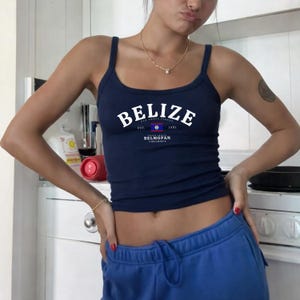 May include: A navy blue ribbed vest top featuring white text that reads BELIZE, EST. 1981, and BELMOPAN, along with a small graphic of the Belizean flag. The garment has a scoop neckline and thin shoulder straps, worn with bright blue sweatpants.