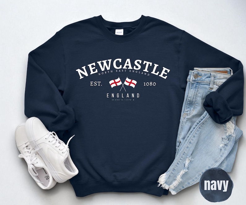Puede incluir: Sudadera azul marino con la palabra "Newcastle" en texto blanco arqueado, dos banderas cruzadas y el texto "England". La sudadera tambi&eacute;n presenta el texto "Est. 1080". Zapatillas blancas y vaqueros azul claro tambi&eacute;n est&aacute;n en la imagen.
