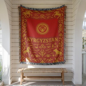 May include: Red and gold woven throw blanket featuring the flag of Kyrgyzstan. The blanket has a central sun emblem, the word "KYRGYZSTAN", and decorative horse and floral motifs. The edges are fringed.