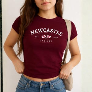 May include: A maroon t-shirt with a white graphic that reads "Newcastle" above "North East England" and "England". The graphic includes two crossed flags and the text "Est. 1080". The shirt has short sleeves and a crew neck.