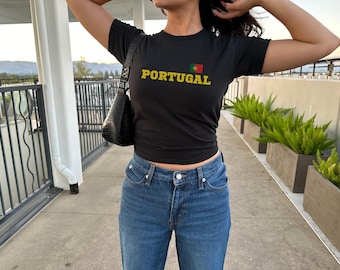 May include: A black t-shirt with the word "PORTUGAL" in yellow lettering and a small Portuguese flag graphic. The person is wearing blue jeans.