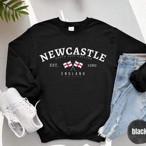 Puede incluir: Sudadera negra con la palabra "Newcastle" en texto blanco arqueado. Debajo, dos banderas cruzadas y las palabras "North East England" y "England". La sudadera tambi&eacute;n tiene el texto "Est. 1080".