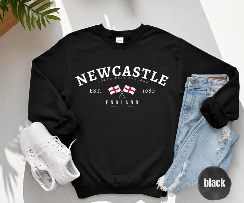 Puede incluir: Sudadera negra con la palabra "Newcastle" en texto blanco arqueado. Debajo, dos banderas cruzadas y las palabras "North East England" y "England". La sudadera tambi&eacute;n tiene el texto "Est. 1080".