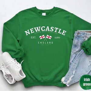 Puede incluir: Sudadera verde irlandesa con la palabra "Newcastle" en texto blanco, con dos banderas cruzadas y el texto "England". La sudadera se combina con vaqueros de lavado claro y zapatillas blancas. El texto "North East England" tambi&eacute;n es visible.