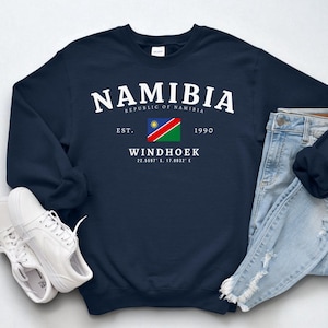 Peut inclure: Sweat-shirt bleu marine avec le mot "NAMIBIA" en blanc. Le drapeau namibien, la ville de Windhoek et les coordonnées sont également présents. Des baskets blanches et un jean bleu sont à côté du sweat-shirt.
