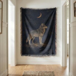 Peut inclure: Un plaid tissé rectangulaire présentant un fond bleu marine foncé avec une illustration centrale d'un loup gris debout. Au-dessus du loup se trouve un croissant de lune doré. La couverture possède un bord à franges décoratif sur les quatre côtés.