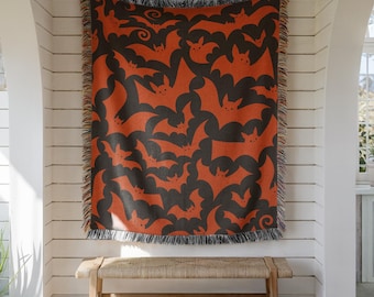 May include: A woven throw blanket featuring a repeating pattern of orange bats on a dark charcoal background. The blanket has a rectangular shape with a decorative fringed edge along the top and bottom, displaying a consistent bat motif throughout.