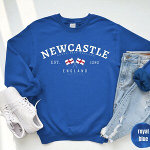 Puede incluir: Sudadera azul real con la palabra "NEWCASTLE" en texto blanco arqueado. Debajo, dos banderas cruzadas y la palabra "ENGLAND". Zapatillas blancas y vaqueros azul claro junto a la sudadera.