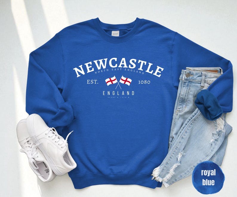 Puede incluir: Sudadera azul real con la palabra "NEWCASTLE" en texto blanco arqueado. Debajo, dos banderas cruzadas y la palabra "ENGLAND". Zapatillas blancas y vaqueros azul claro junto a la sudadera.