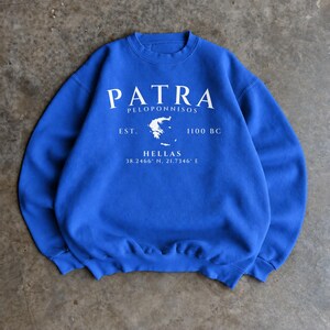 May include: A royal blue crewneck sweatshirt featuring white screen-printed text that reads PATRA PELOPONNISOS EST. 1100 BC HELLAS 38.2466° N, 21.7346° E, centred around a small white graphic outline of the map of Greece.
