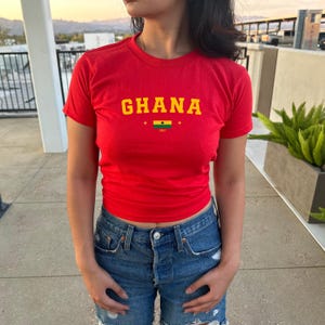 K&ouml;nnte beinhalten: Ein rotes T-Shirt mit dem Wort "GHANA" in gelben Buchstaben und einer kleinen ghanaischen Flagge mit dem Text "1957" darunter.