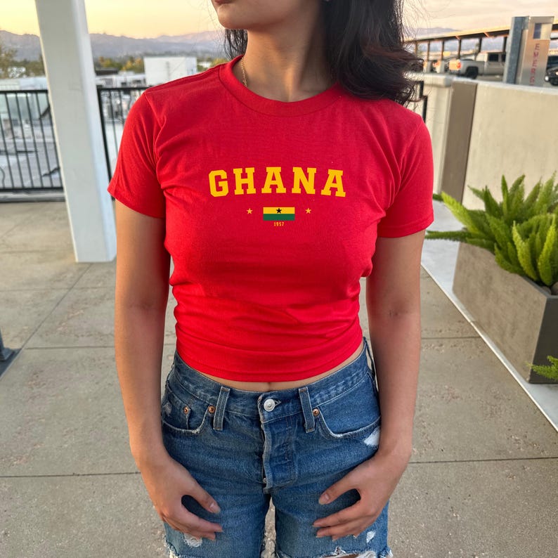 K&ouml;nnte beinhalten: Ein rotes T-Shirt mit dem Wort "GHANA" in gelben Buchstaben und einer kleinen ghanaischen Flagge mit dem Text "1957" darunter.