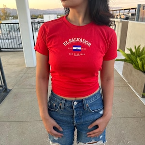 Może przedstawiać: Czerwony T-shirt z białym nadrukiem flagi Salwadoru i tekstem "El Salvador" z napisami "Est. 1524" i "San Salvador" poniżej.