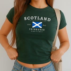 May include: Dark green crop top with the word "SCOTLAND" in white arched text. Below is a white and blue flag, the text "EDINBURGH" and coordinates. The text "In My DEFENS GOD ME DEFEND" is above the word "SCOTLAND".