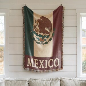 May include: A woven throw blanket featuring the Mexican flag design, with green, white, and red vertical stripes. The center displays the Mexican coat of arms, and the word "MEXICO" is at the bottom. The blanket has fringed edges and is hanging on a white wall.