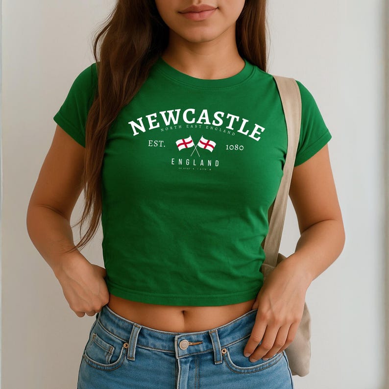May include: A green crop top with the word "NEWCASTLE" in white arched across the chest. Below the word are two crossed flags and the words "EST. 1080 ENGLAND". The shirt has short sleeves and is paired with blue jeans.