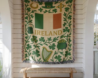 May include: A woven throw blanket featuring the Irish flag in green, white, and orange with the word IRELAND printed in gold. The design includes green shamrocks, a leprechaun hat, a horseshoe, a Celtic cross, a harp, and a pot of gold.