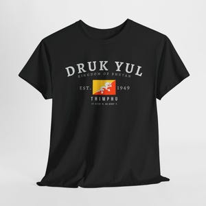 May include: Black t-shirt with white text that reads "Druk Yul Kingdom of Bhutan Est. 1949 Thimphu 27.472° N, 89.639° E". A small Bhutanese flag is printed on the shirt.