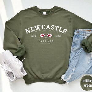 Puede incluir: Una sudadera verde oliva con la palabra "Newcastle" en texto blanco arqueado. Debajo, dos banderas cruzadas y la palabra "England". Un par de zapatillas blancas y unos vaqueros azules est&aacute;n al lado de la sudadera.
