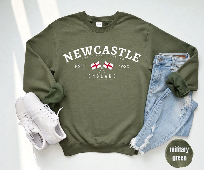 Puede incluir: Una sudadera verde oliva con la palabra "Newcastle" en texto blanco arqueado. Debajo, dos banderas cruzadas y la palabra "England". Un par de zapatillas blancas y unos vaqueros azules est&aacute;n al lado de la sudadera.