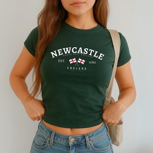 May include: Dark green t-shirt with the word "NEWCASTLE" in white arched text. Below the text are two crossed flags and the word "ENGLAND". The shirt also has the text "NORTH EAST ENGLAND" and "EST. 1080".
