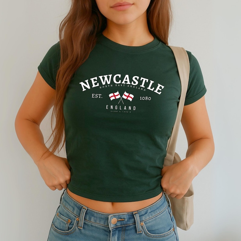 May include: Dark green t-shirt with the word "NEWCASTLE" in white arched text. Below the text are two crossed flags and the word "ENGLAND". The shirt also has the text "NORTH EAST ENGLAND" and "EST. 1080".