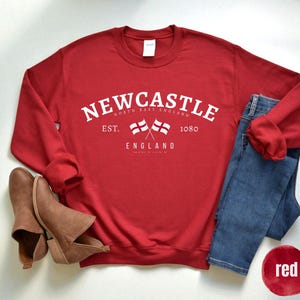 Puede incluir: Una sudadera roja con el texto blanco "NEWCASTLE NORTH EAST ENGLAND", dos banderas cruzadas y el a&ntilde;o "1080". La imagen incluye vaqueros azules, botines marrones y un c&iacute;rculo rojo con la palabra "red".