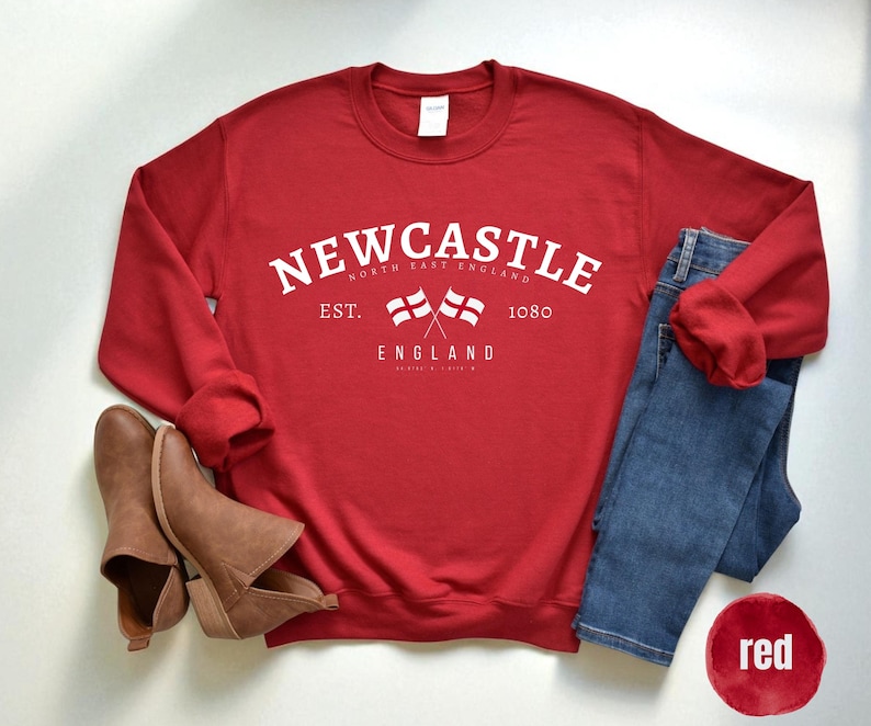 Puede incluir: Una sudadera roja con el texto blanco "NEWCASTLE NORTH EAST ENGLAND", dos banderas cruzadas y el a&ntilde;o "1080". La imagen incluye vaqueros azules, botines marrones y un c&iacute;rculo rojo con la palabra "red".