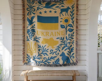 May include: A woven throw blanket featuring a beige background with blue and yellow patterns. The design includes the Ukrainian flag, the word UKRAINE, a map of the country, a trident symbol, sunflowers, birds, and foliage with fringed edges.