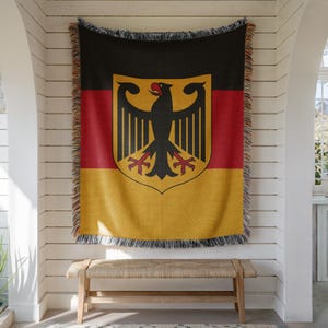 May include: A woven throw blanket featuring the German flag with horizontal black, red, and gold stripes. A large black eagle emblem with a red beak and red talons is centered on the blanket. The edges are finished with a multi-colored fringe.