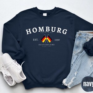 Op de afbeelding: Marineblauw sweatshirt met het woord "Homburg" in het wit, samen met de tekst "Saarland" en "Deutschland". Het sweatshirt heeft twee gekruiste Duitse vlaggen en het jaar "1330". Witte sneakers en een blauwe jeans zijn ook zichtbaar.